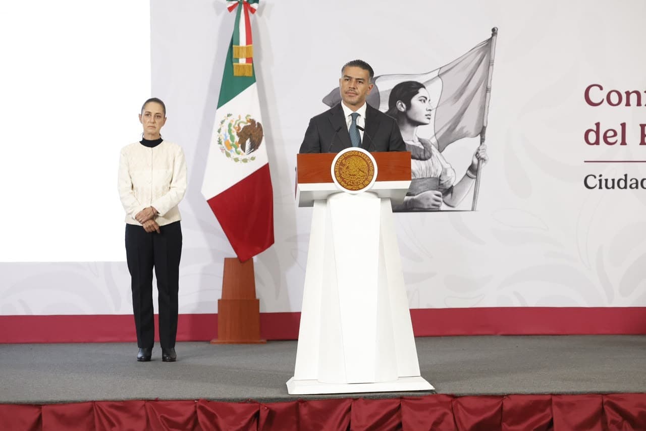 Omar García Harfuch, secretario de Seguridad, ofrece una conferencia en Querétaro junto a una mujer, con la bandera mexicana de fondo.
