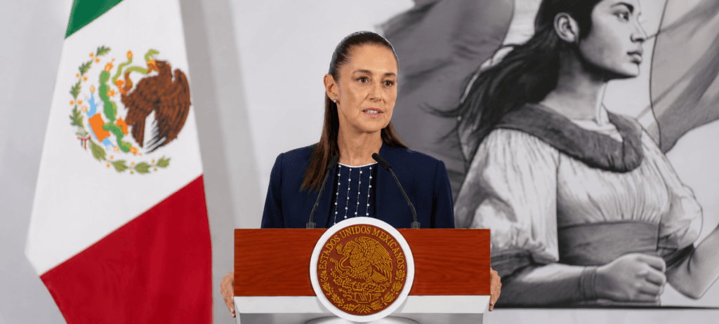 Claudia Sheinbaum durante su conferencia matutina, hablando sobre la relación con Estados Unidos y el T-MEC, con la bandera de México de fondo.