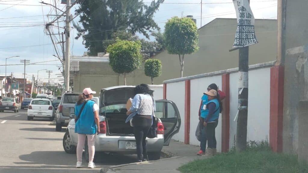 Encuestadores de Morena en Tlaxcala realizan su trabajo en la calle, con un vehículo estacionado y un grupo de personas en la acera.